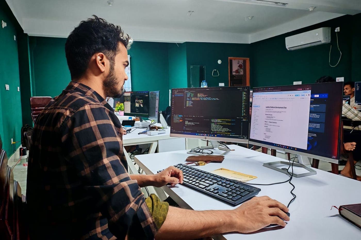 Developer coding at a dual-monitor desk in a green-walled office