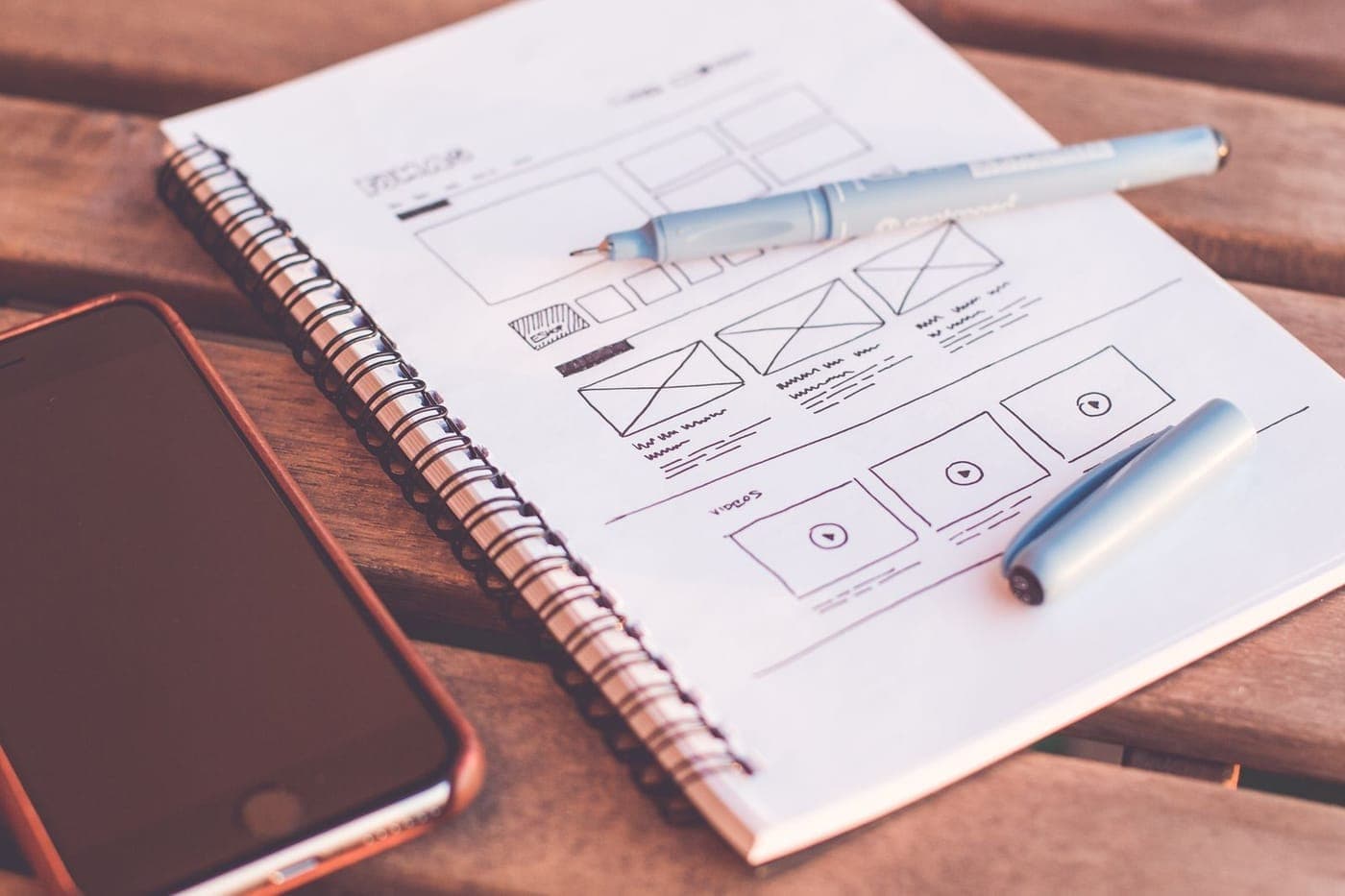 Hand-drawn UI wireframe sketch in a spiral notebook with pens on a warm wooden desk