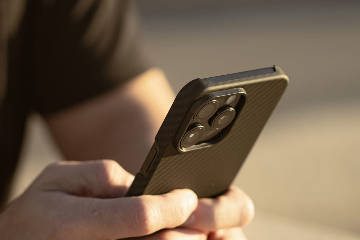 Hands holding a modern smartphone with a carbon-fiber case in warm golden light