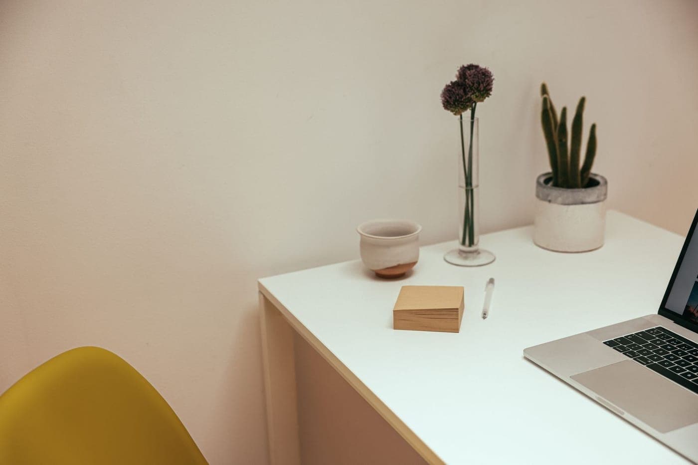 Calm minimal white desk with a laptop, notepad, plant and a yellow chair in warm light