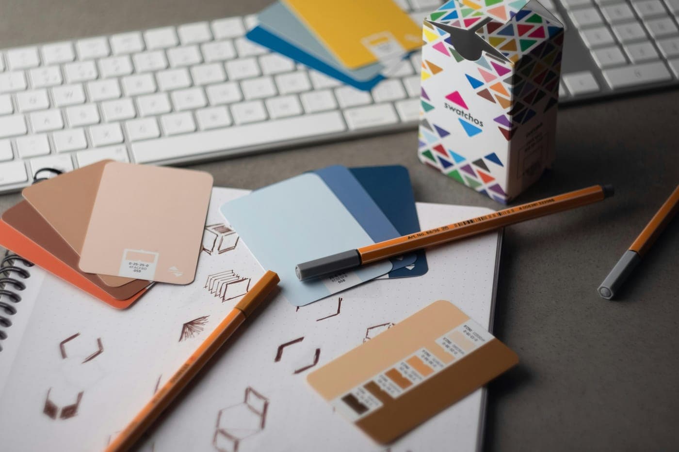 Designer's desk with color swatch cards, a pen, and interface sketches in a dotted notebook beside a keyboard