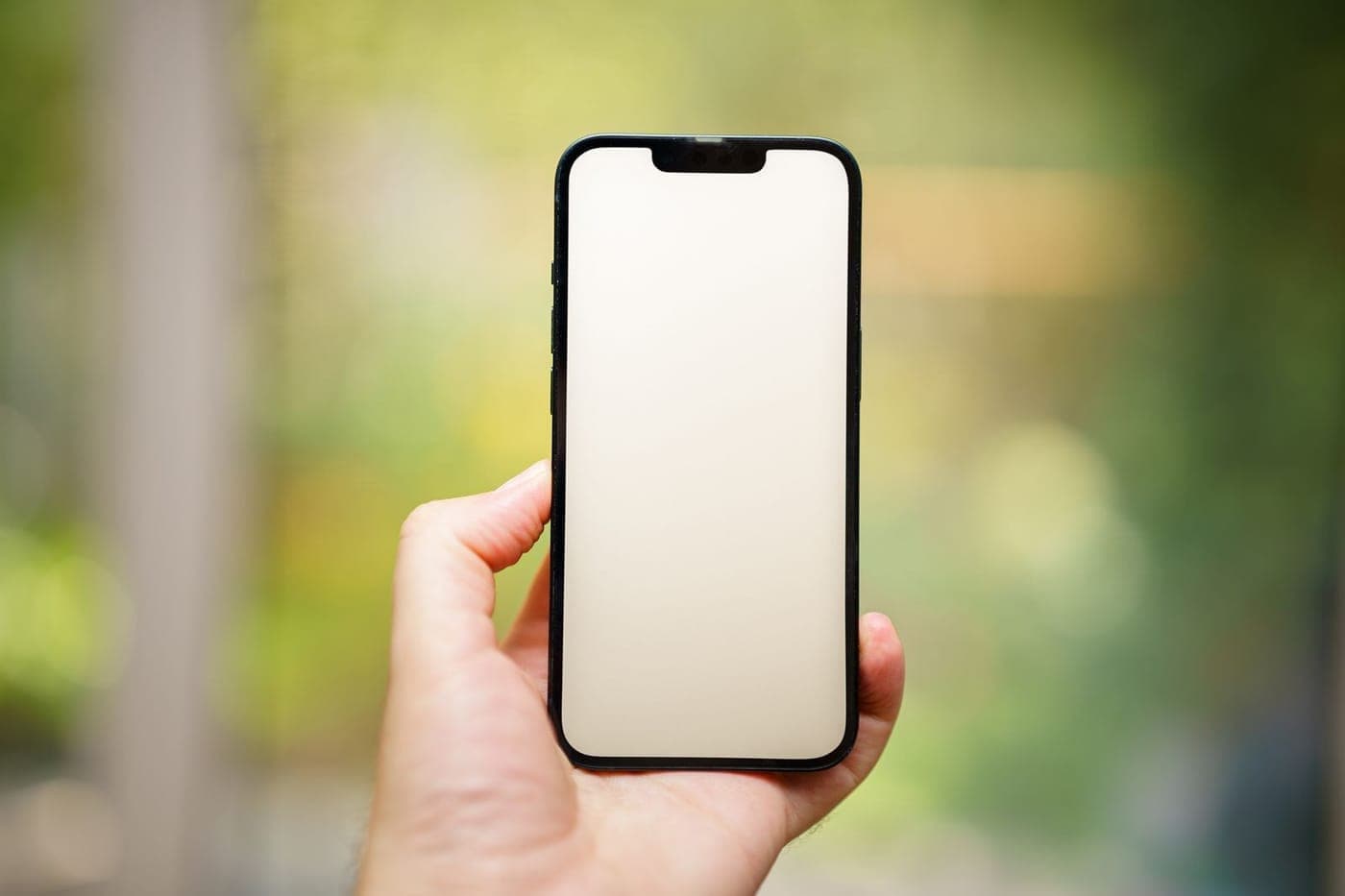 A modern iPhone with a blank cream screen held in hand against a soft green bokeh background