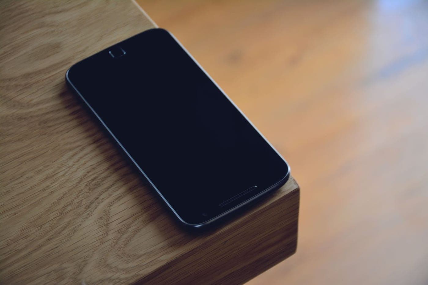 A black smartphone resting on the edge of a wooden table in warm light with shallow depth of field