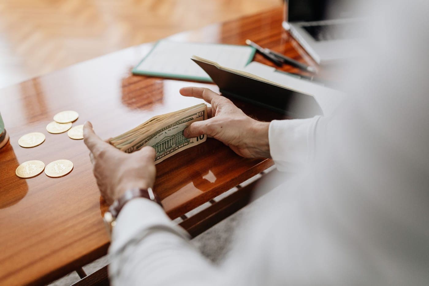 Hands counting cash and gold coins beside a notebook and laptop on a warm wooden desk