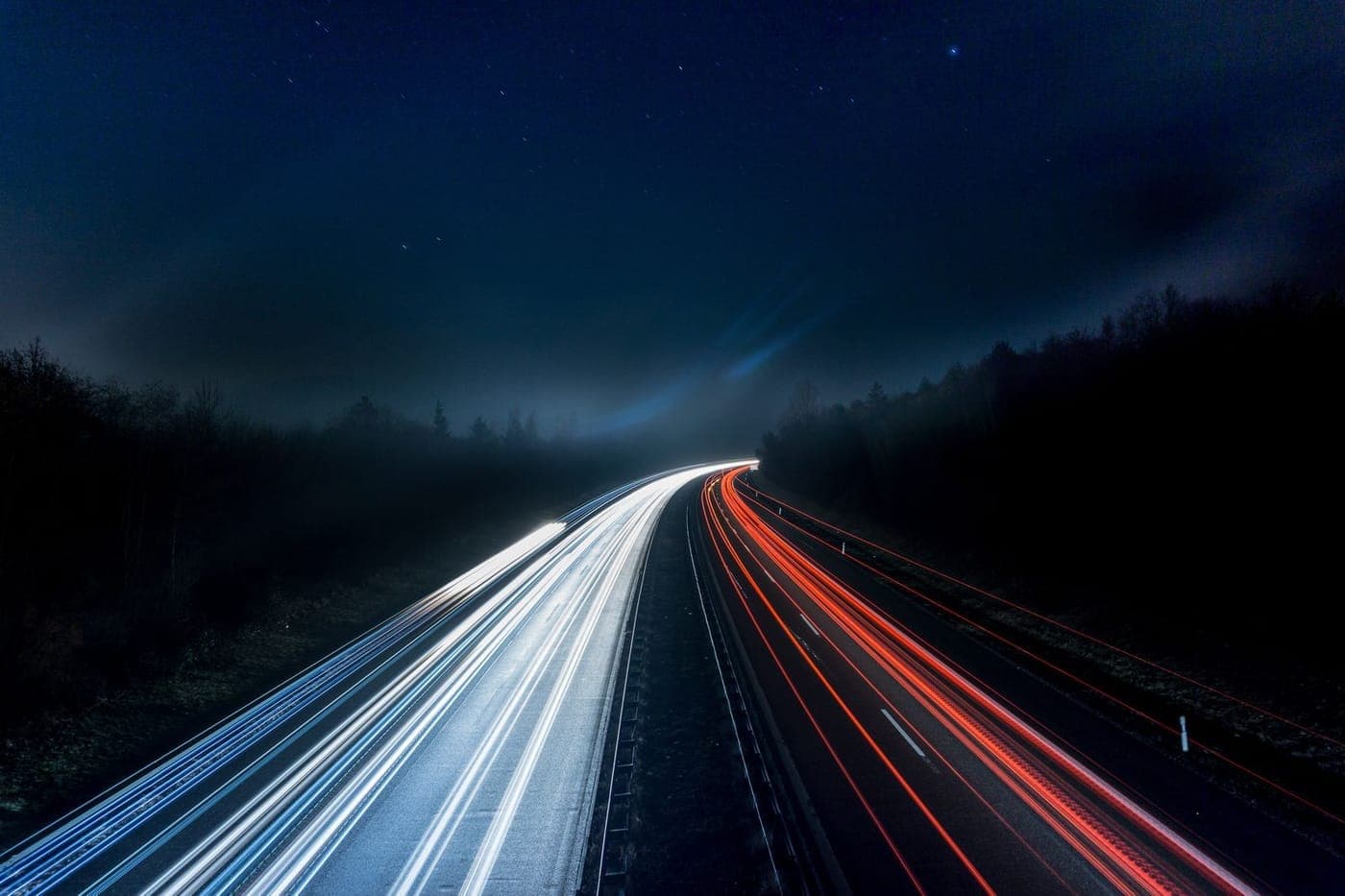 Long-exposure blue and red light trails sweeping along a curved highway under a night sky